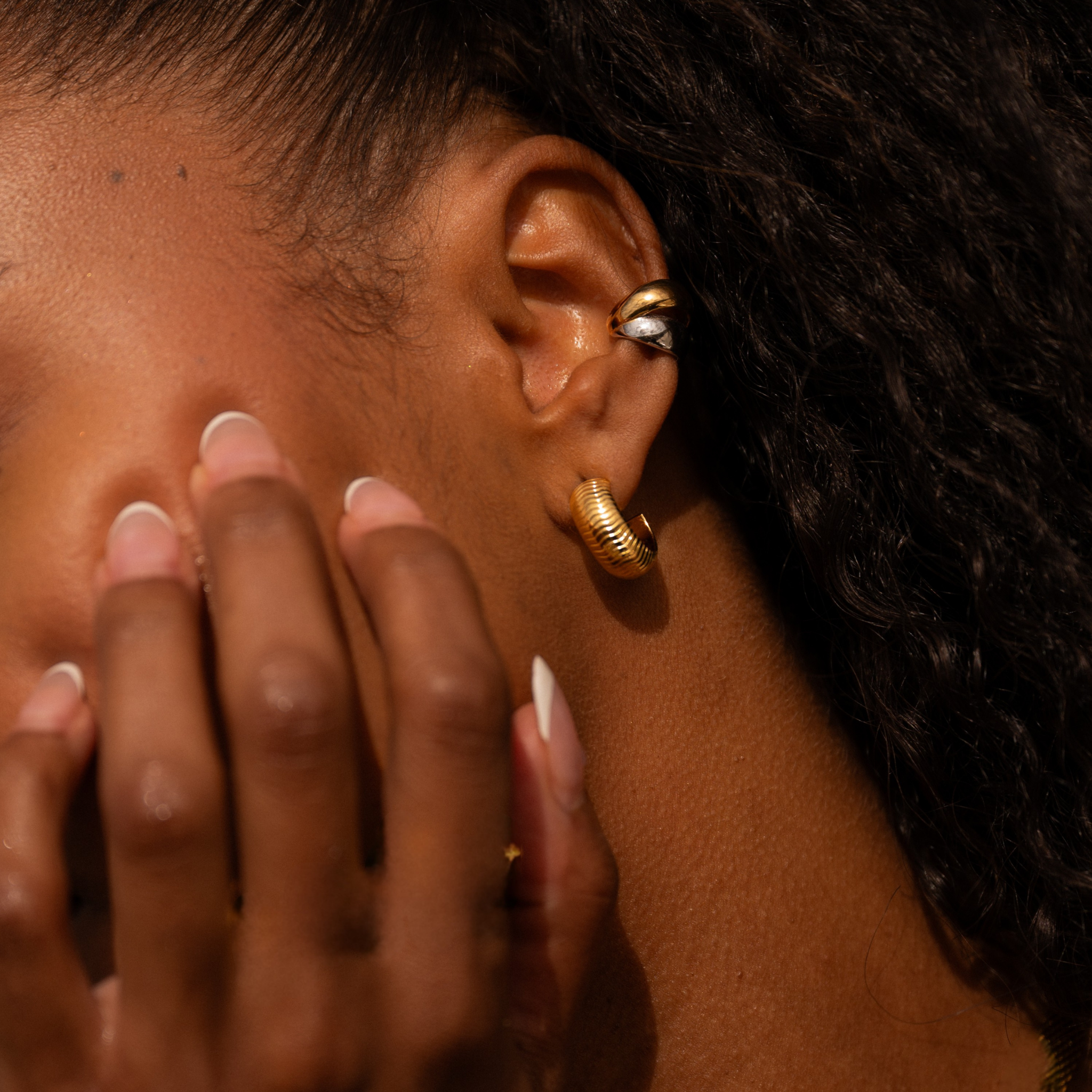 Close-up of an ear with gold earrings with wave engraving and a hand touching the cheek. Waterproof